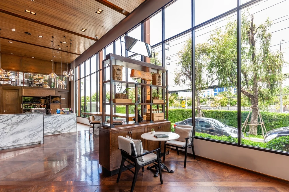 Modern cafe interior with large windows, wooden furniture, marble countertop, and indoor greenery.