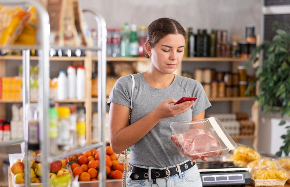 Young female shopper scanning qr code for packaged raw pork meat in grocery store.